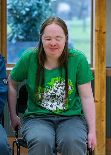A woman is sat down on a chair in the Pimlott building. She is smiling with her eyes closed, and wearing a green t-shirt with sheep on it.