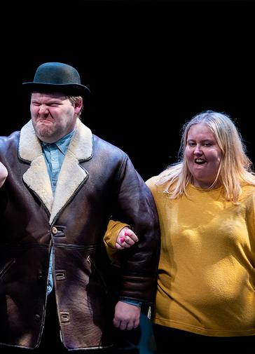 Two people stand on stage, with their arms linked. They both have big expressions on their faces. On the left is a man in a leather jacket and a black bowler hat scrunches his face up angrily. On the right, a woman in a yellow jumper who is smiling.