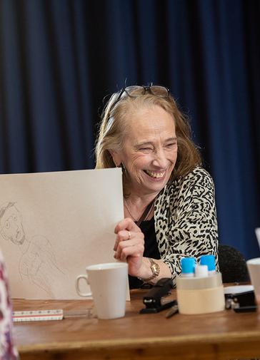 A group of people are in a drawing workshop. A lady is smiling holding up her drawing.