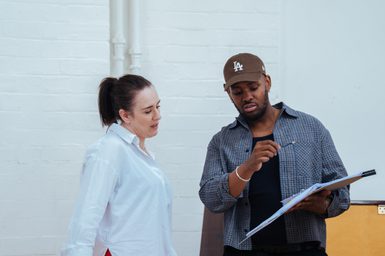A man and woman in a rehearsal room. They are both reading from a document he is holding.