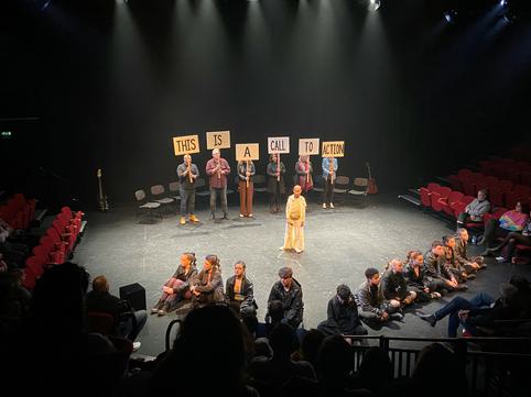 A group of young performers in the Minerva Theatre, sat on the floor looking out at the audience wearing black clothes. One performer in white robes stands centre stage. At the back of the stage, six older people hold up signs saying 'This Is A Call To Action'.