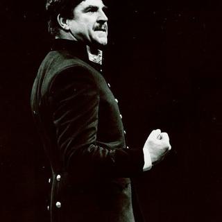 An actor stands side with a clenched fist, wearing a hat and military uniform