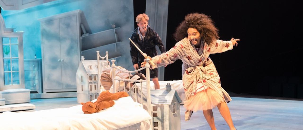 Production photo from Peter Pan. A young girl with dark curly hair wearing a grey dressing gown over a puffy skirt with grey slippers is holding onto a bed frame. There is a teddy lying on the bed. She looks to be running around it and is smiling. Behind her is a smaller boy in a navy blue dressing gown who is looking at her and smiling. There are small white houses on the floor around them and a white window, wardrobe, drawers and shelves can be seen in the background.