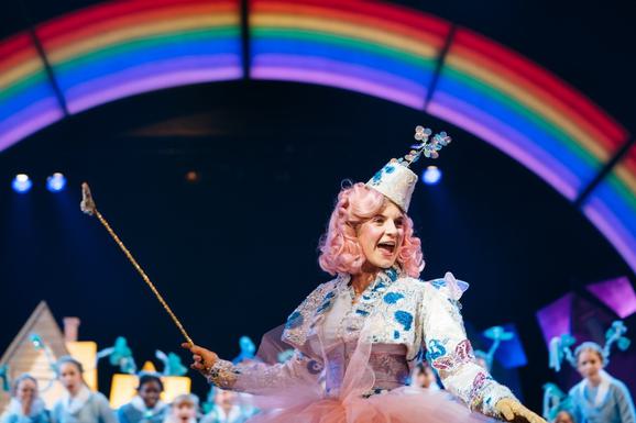 Production photo from The Wizard of Oz. A girl with pink curly hair wearing a silver and blue embellished jacket and matching hat holds a long, gold, sparkling wand. She looks off to the right with her mouth open, like she is singing. In the background, there is a rainbow in lights. Other young performers are out of focus, looking at her in awe.