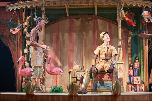 Production photo from Pinocchio. Two young male performers are on stage, surrounded by wooden toys. One sits on a colourful chest of drawers and is wearing a brown costume that makes it look like he is made of wood. He looks off to the right with a joyous expression. The other performer stands to the left, looking at him, wearing a brown apron. He has grey hair, exaggerated sideburns and glasses and looks concerned or shocked.