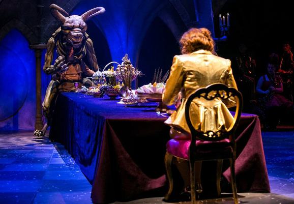 Production photo from Beauty and the Beast. There is a long table on stage with a velvet tablecloth and an impressive banquet spread. The lighting makes everything appear purple. At one end of the table sits a woman with her back to the camera, wearing a golden blazer with her hair tied up. At the other, there is a huge beast puppet with scary horns and claws, looking menacing.