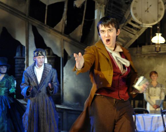 Production photo from A Christmas Carol. A boy with dark hair looks up with his hand outstretched and his mouth open like he is speaking or singing. He is wearing a red waistcoat, brown trousers and a long brown jacket. In the background there is another boy with a long black coat and black hat, looking at him, and two other performers who are out of focus.