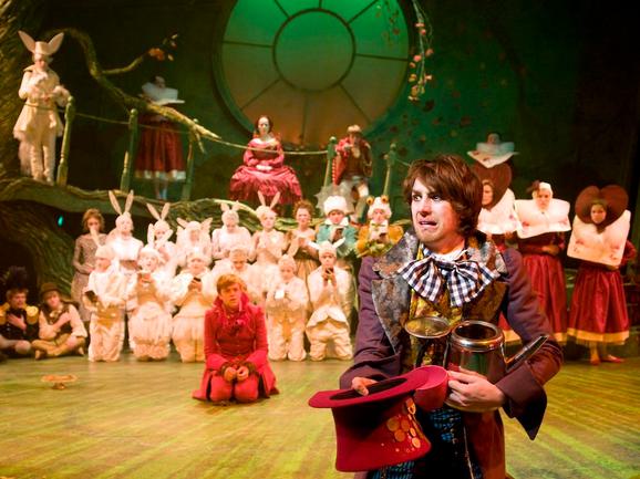Production photo from Alice in Wonderland. A man with brown hair and stubble looks off to the right with an alarmed expression. He is holding a red top hat and wearing a purple coat and checked bow tie. In the background an ensemble of other performers can be seen, looking at him.