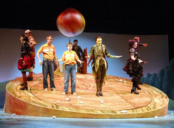 Production photo from James and the Giant Peach. Eight performers stand on a wooden platform with a big peach hanging above them. They are singing and holding their arms out.