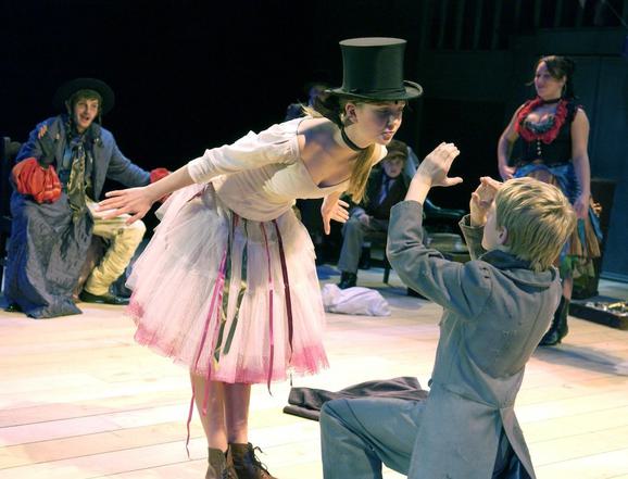 Production photo from Oliver. A girl wearing a white top, white and pink poofy skirt and a black top hat bends over a boy in a grey suit with blonde hair, who is down on one knee. She looks down at him and he holds his arms up as though looking at her through a telescope.