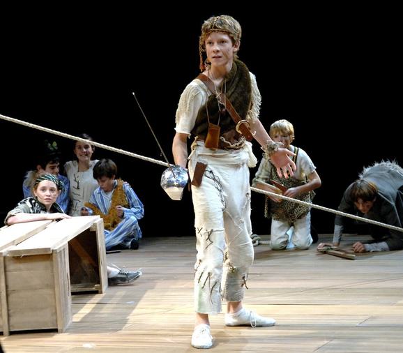 Production photo from Peter Pan. A young boy wears ragged white trousers and a white shirt with brown fur tied over his shoulder. He holds a silver sword and looks slightly off to the left with a serious expression. Other young performers crouch on the stage around him.