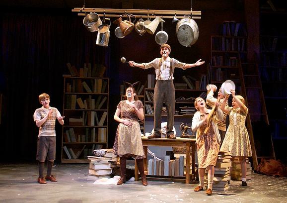 Production photo from The Lion, The Witch and The Wardrobe. A young boy with his face painted like an animal stands on a wooden table. A girl stands below him with the same face paint and four other young performers walk around them, banging kitchen pots like they are celebrating.