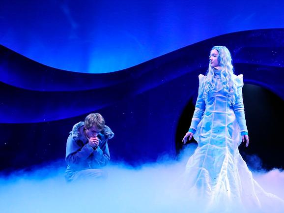 Production photo from The Snow Queen. The stage is lit in blue and covered with smoke. A young male performer crouches and looks down. A female performer stands to the right, looking down at him. She has long silver hair and wears an impressive blue gown.