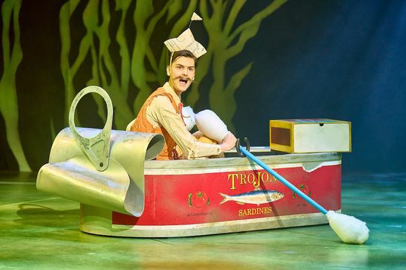 Production photo from The Wind in the Willows. A male performer sits in a boat that looks like a sardine tin, holding a cotton bud oar. He has a dramatic brown moustache and wears a paper boat as a hat.