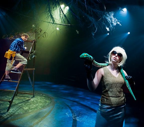 Production photo from The Witches. A female performer wears a blonde wig, sunglasses, a sparkly gold top and black gloves. She holds a green snake around her neck. In the background, a young boy climbs a ladder and looks at her inquisitively.