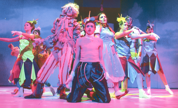 Production photo from The Wizard of Oz. A group of young performers wearing colourful costumes are huddled together with their arms outstretched, dancing. A young performer sits on his knees at the front of the group. They are all lit in bright pink.