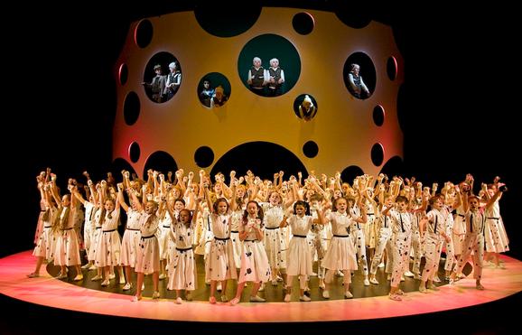 Production photo from The Hundred and One Dalmatians. The stage is full of young performers dressed as Dalmatians with their arms in the air, singing.