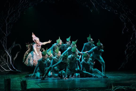 Production photo from Sleeping Beauty. The stage is lit in dark green. A group of young performers wearing green costumes and hats are huddled together, looking menacing. A female performer stands behind them, wearing a big dress and pink pointy hat.
