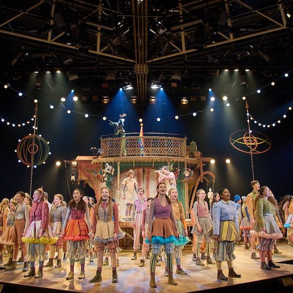 Production photo from Pinocchio. A group of young performers wearing colourful costumes all stand proudly around the stage, looking out to the audience and singing.