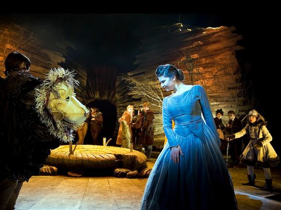 Production photo from The Lion, The Witch and The Wardrobe. On the left is the head of a lion puppet. On the right, is a girl wearing a blue velvet dress with her dark hair tied up, bowing at the lion.