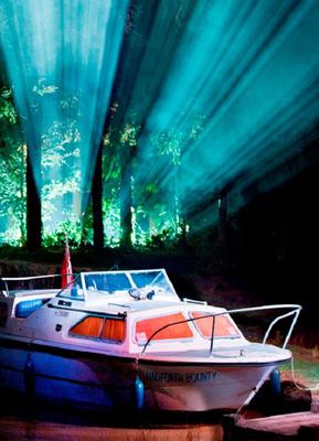 A white boat sits at a riverbank. Light streams in blue and white through trees behind it.