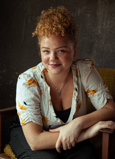 A woman with short brown curly hair swept on top of her head sits with her arms crossed, smiling at the camera. She wears a white blouse patterned with yellow and orange flowers and black flecks, over a black top and trousers, and a silver pendant necklace.