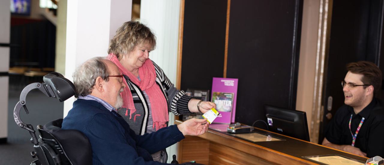 A bearded male wheelchair user, accompanied by a woman wearing a pink scarf, smile as they receive a ticket from a cheerful Box Office assistant