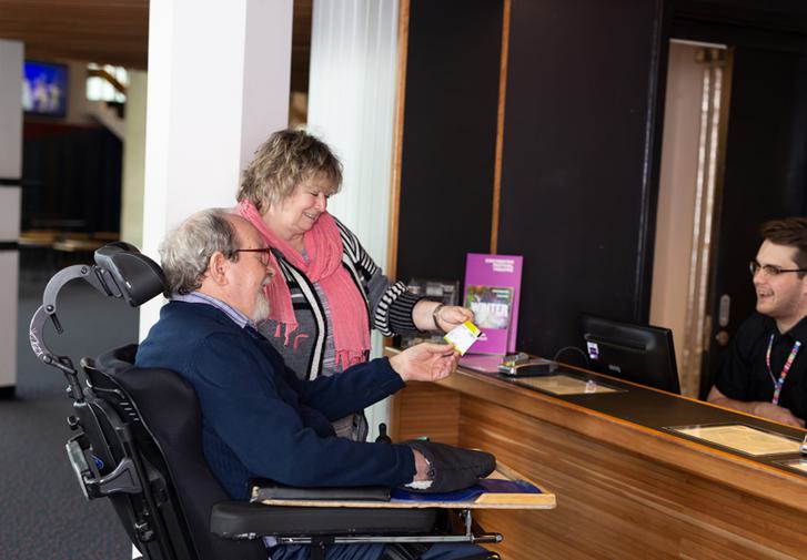 A bearded male wheelchair user, accompanied by a woman wearing a pink scarf, smile as they receive a ticket from a cheerful Box Office assistant