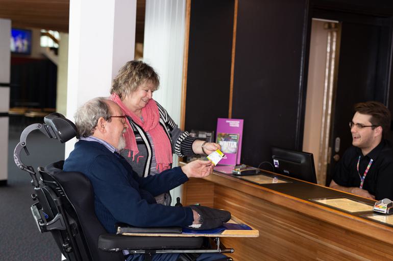 A bearded male wheelchair user, accompanied by a woman wearing a pink scarf, smile as they receive a ticket from a cheerful Box Office assistant