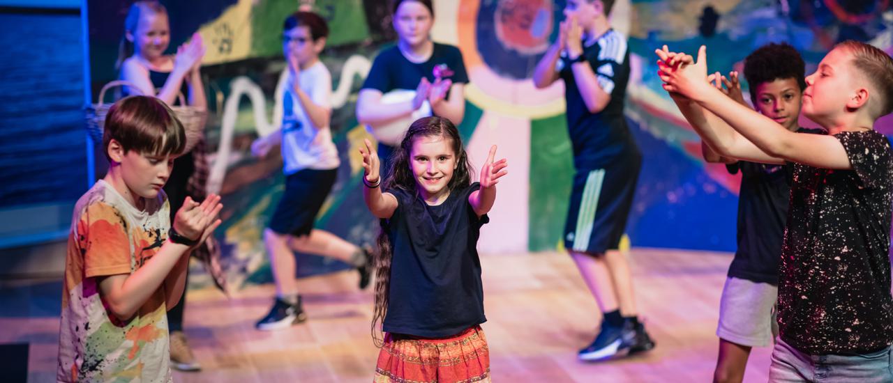 A group of young people of varying ages performing on the Minerva Theatre stage against a colourful backdrop they have painted themselves.