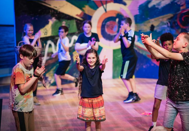 A group of young people of varying ages performing on the Minerva Theatre stage against a colourful backdrop they have painted themselves.
