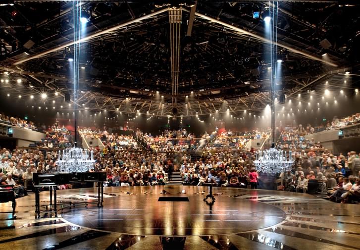 A view of the Festival Theatre auditorium, taken from the back of the stage and looking out into the audience, seated in an amphitheatre. The stage has a dull gold parquet floor with star-shaped panels reflecting the light; there is a harpsichord on the left and two crystal chandeliers which have been lowered to a few feet from the floor. The audience are illuminated by the house lights which are suspended from a central, hexagonal truss.