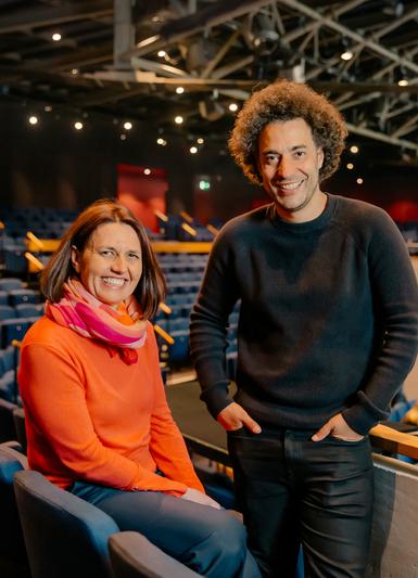 Kathy Bourne and Justin Audibert are pictured in the Festival Theatre auditorium, smiling broadly towards the camera. Kathy sits, wearing an orange jumper, pink and orange scarf and blue trousers, and has shoulder length brown hair. Justin wears black and has curly brown hair, standing with his hands in his pockets