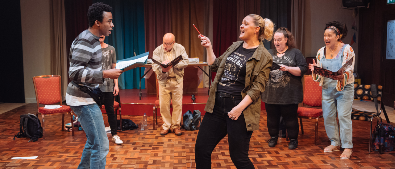 A group of performers are stood in a semi-circle, singing gloriously while reading from folders. In the centre, a woman with blonde hair is using a pencil like a conductor's baton. They are in a dated rehearsal room, with retro wooden floors, colourful curtains and fairy lights.
