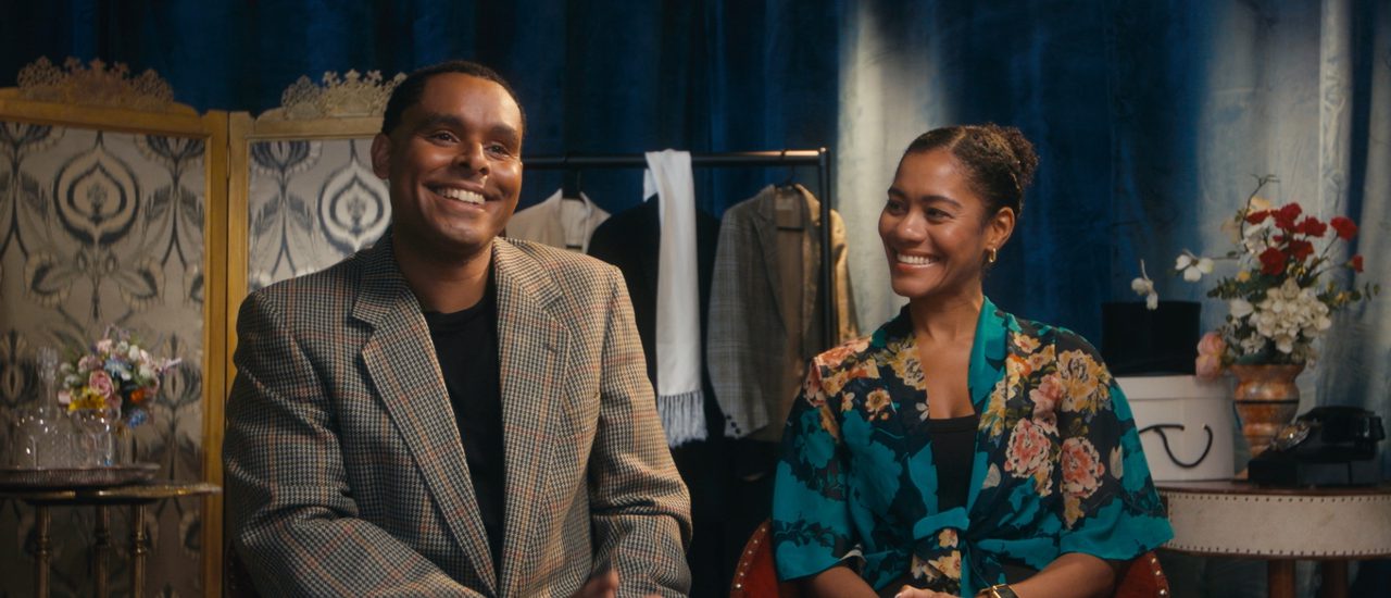 Phillip Attmore and Lucy St. Louis are sat smartly dressed and smiling at each other. In the background is a blue curtain and an ecclectic mix of show props and costumes.