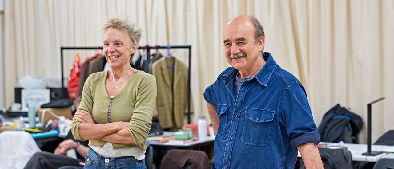 A woman with spiky blonde hair is stood with her arms crossed. Next to her is a man with a grey moustache leaning against a table. They are both smiling.