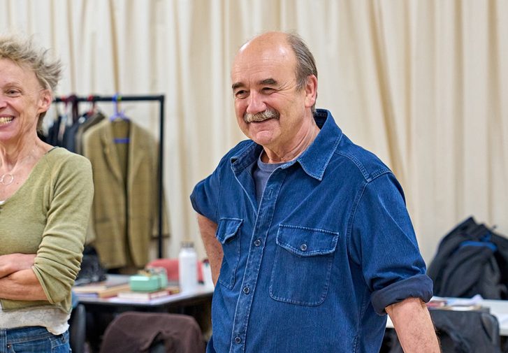 A woman with spiky blonde hair is stood with her arms crossed. Next to her is a man with a grey moustache leaning against a table. They are both smiling.