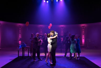 A couple kiss passionately at a dance. Others stand around them, dancing and socialising. The room is lit in shades of purple and pink.