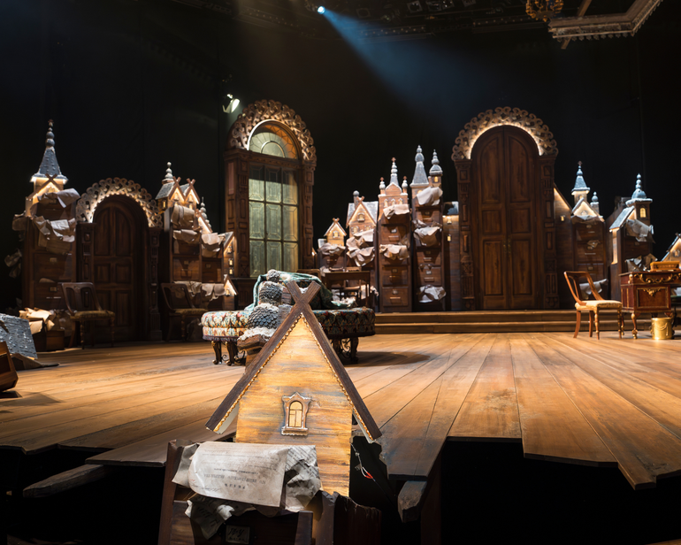 The stage set for The Government Inspector with atmospheric warm lighting. In the foreground is a small wooden house. The floor of the stage is covered with wooden floorboards. In the centre is a lavish circular couch. Around the stage are doors, windows and lots of wooden filing cabinets, decorated with little wooden houses on the top of each one, with papers spilling out from them. On the far right is a wooden desk and chair.
