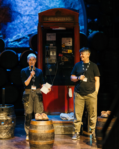 Two members of the crew stand on stage amongst the set: there are barrels littered around the stage floor and a telephone box stands behind them, with atmospheric blue lighting and a pile of barrels in the background.