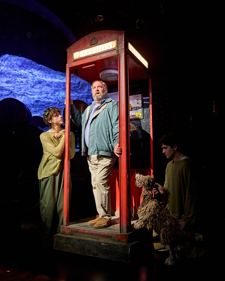 An older man wearing casual clothes is stood inside a worn-down red telephone box. His hands are clasping the doorframe, and he is looking slightly worried. To his right, a young man dressed in an almost threadbare green jumper with a crown of leaves in his hair is gazing up at him inquisitively. On the other side, a shaggy dog puppet operated by a puppeteer is also looking at the man in the phone box.