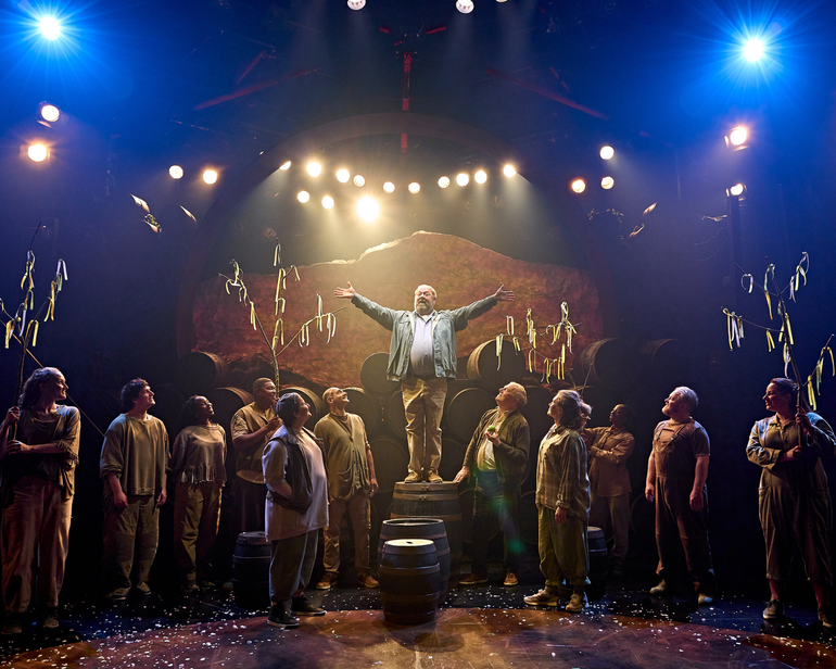 Many characters are gazing wondrously at a man is standing atop a barrel in the centre of the stage. He has his arms wide open. Some of the ensemble are holding tree branches with ribbons tied to them. There are many barrels stacked in the background and others placed amongst the group of people.