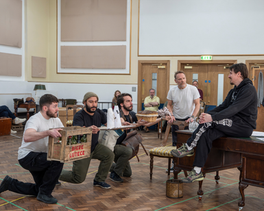 Performers wearing casual clothes rehearse in a studio. Three performers crouch on one knee on the floor, in a line. They each hold out a different object: a crate of beer, a basket of bread and a tray with something balancing on it. They look concerned. Opposite them, sitting on a dark wooden vintage desk, another performer looks down at them, leaning back and looking much more relaxed than the others. A fifth performer hovers behind him, leaning on the back of a chair, watching attentively. In the background, other members of the company watch on.