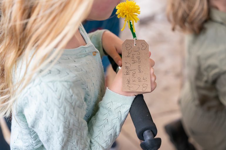 A young girl holds a woollen dandelion flower with a brown paper tag attached, which reads: 'be happy be kind you no you can do it'.