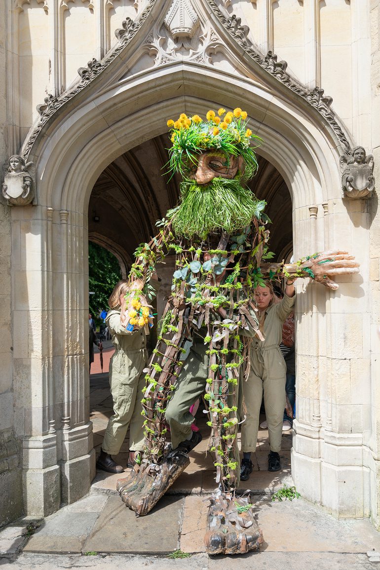 A tall puppet emerging from a stone archway. The puppet is woven from natural materials, with wooden arms and legs, a green, grassy beard and dandelion flowers for hair.