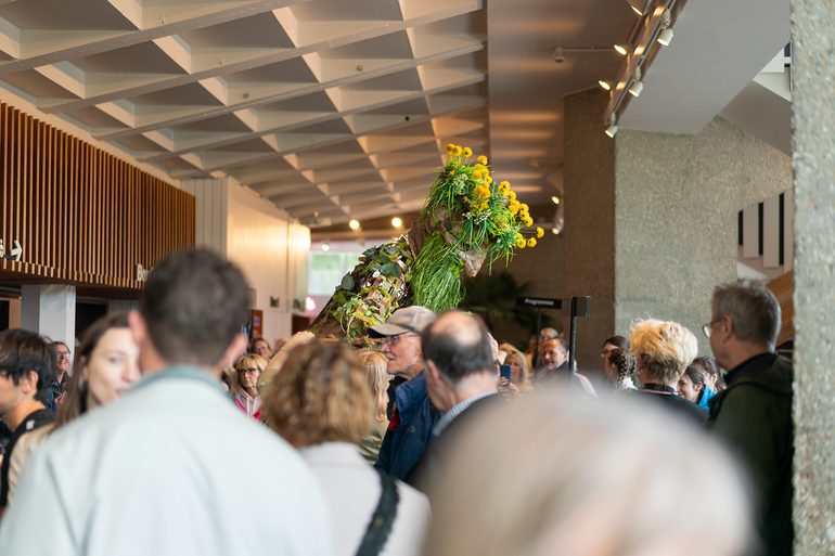 A huge puppet stands in a crowded foyer space. The puppet has a green, grassy beard, oversized, wooden features and dandelion flowers for hair.