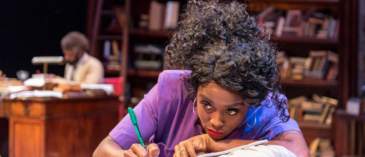Production photograph of Lashana Lynch in Educating Rita. She sits at a wooden desk, wearing a purple short-sleeved blouse with her black curly hair tied up and big hoop earrings. She leans on one hand and holds a green pen in the other, looking out into the distance with a vacant expression. A man can be seen sitting at another desk in the background, out of focus, surrounded by wooden bookshelves full of books.