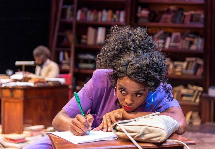Production photograph of Lashana Lynch in Educating Rita. She sits at a wooden desk, wearing a purple short-sleeved blouse with her black curly hair tied up and big hoop earrings. She leans on one hand and holds a green pen in the other, looking out into the distance with a vacant expression. A man can be seen sitting at another desk in the background, out of focus, surrounded by wooden bookshelves full of books.