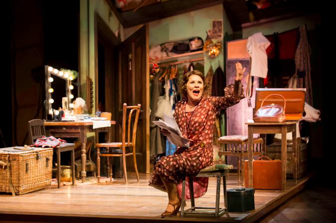 Production photograph from Gypsy starring Imelda Staunton, who sits centre stage on a low wooden stool, wearing a brown dress with pink and orange spots, brown shoes and a necklace. She holds a bunch of papers in one hand and wanes out to the audience with the other. She looks up slightly to the left of the camera, with her mouth open singing or speaking passionately. In the background is a dressing room with brown suitcases, a clothes rail and a dressing table with a lit mirror and wooden chair.
