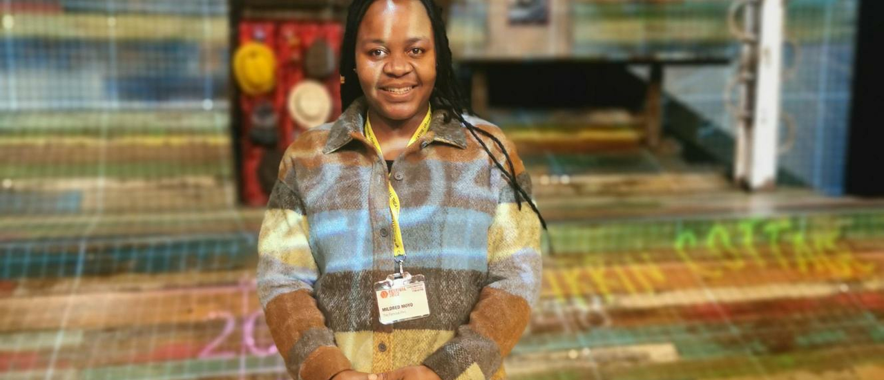 A young woman wearing a checked shirt and a CFT lanyard around her neck stands in the middle of the colourful set of The Famous Five